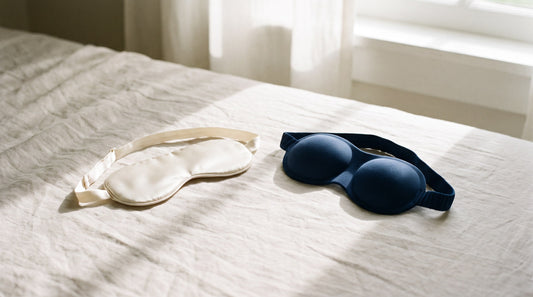 A flat ivory silk sleep mask and a contoured navy sleep mask laid side by side on linen